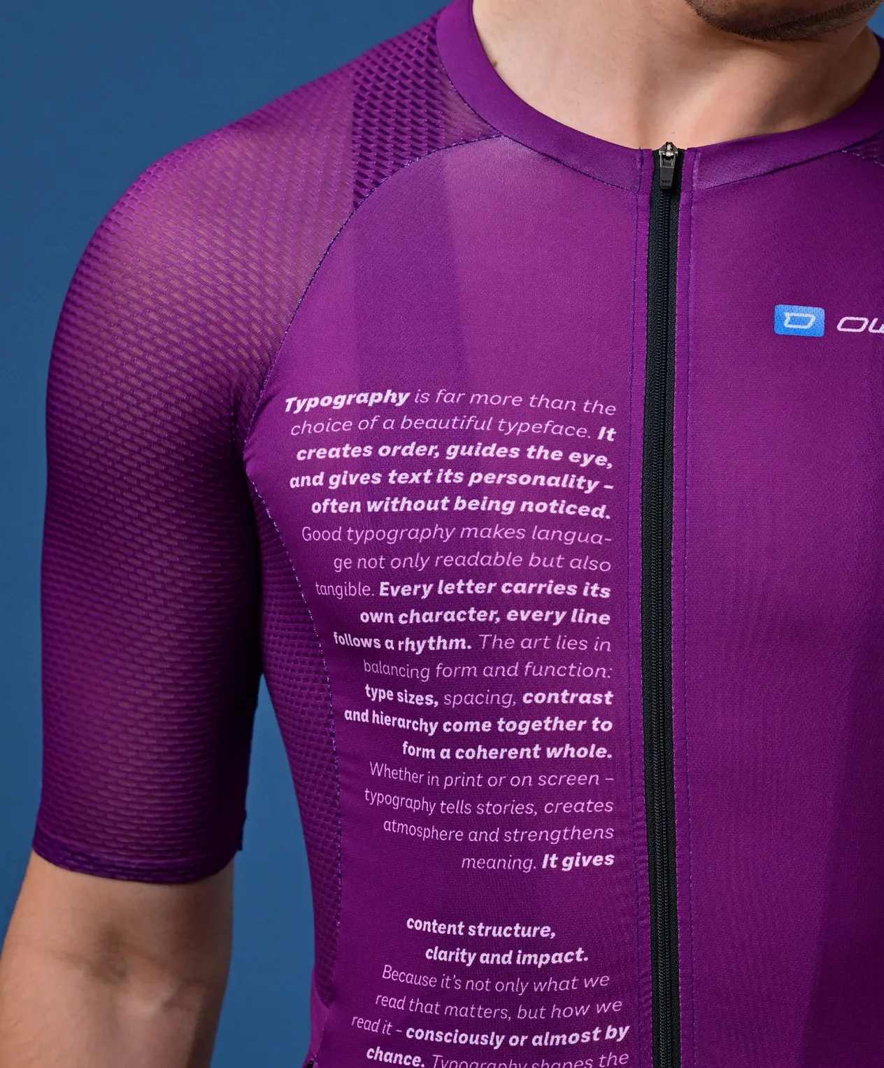 Een man draagt een paars, korte mouwen sportshirt met een tekst over typografie op de borst en een klein logo op de schouder.