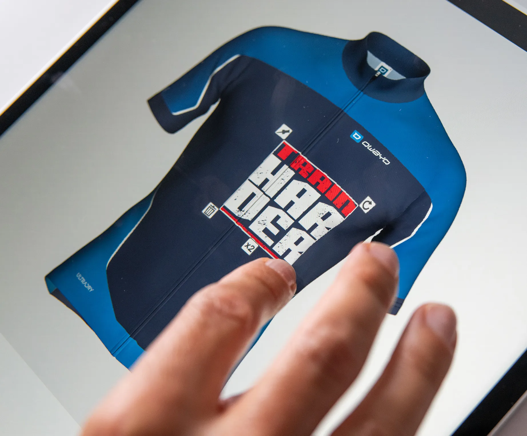 Een hand raakt een scherm aan waarop een aangepast donkerblauw en blauw sportshirt met de tekst 'TRAIN HARDER' en het owayo-logo wordt weergegeven.