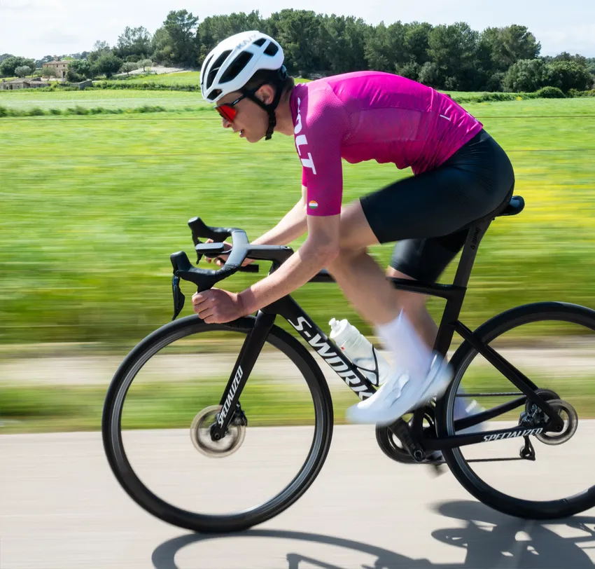 Een fietser in een roze trui en zwarte korte broek rijdt op een weg door een groene landschap, waarbij de bewegingsonduidelijkheid de snelheid benadrukt.