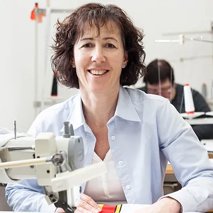 Een vrouw glimlacht naar de camera terwijl ze aan een naaimachine werkt in een werkplaats, met een andere persoon op de achtergrond die ook aan een naaimachine werkt.