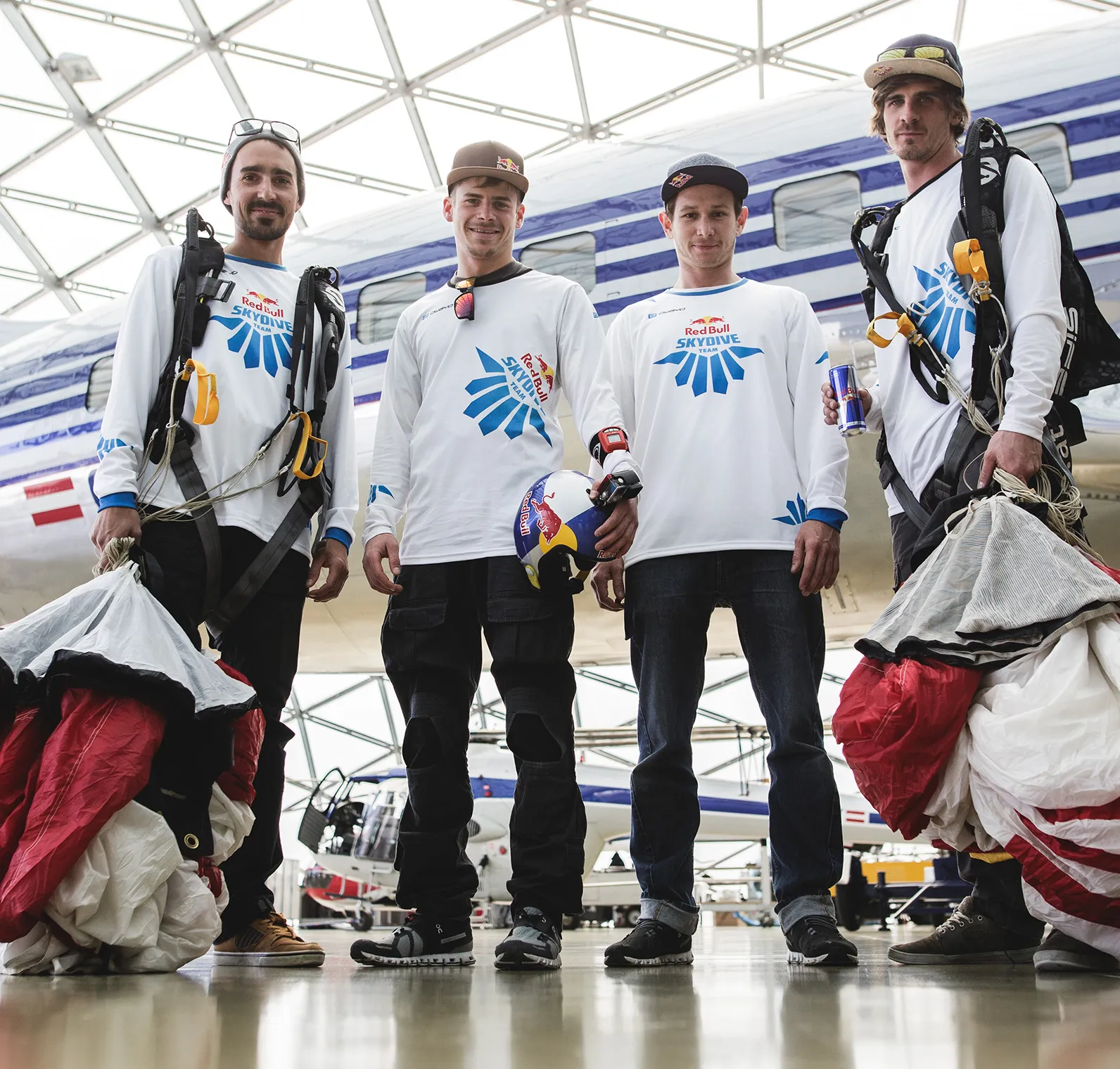 Vier mannen in witte, lange mouwen sportshirts met het logo van de Red Bull Skydive Team staan in een hangar voor een vliegtuig.