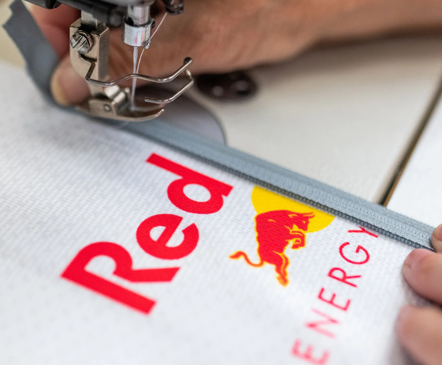 Een naaimachine naait een grijze rits aan een wit sportkledingstuk met het Red Bull-logo.