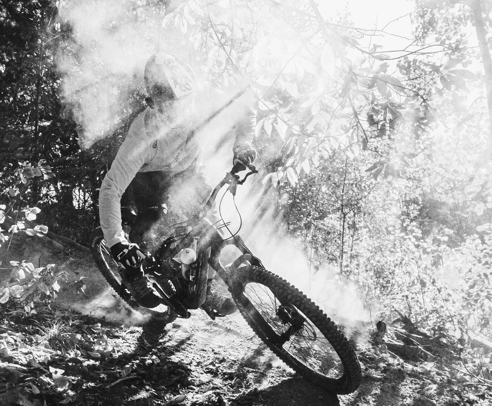 Een mountainbiker in beweging op een bospad, omringd door stof en lichtstralen, draagt een lang mouw shirt van owayo.