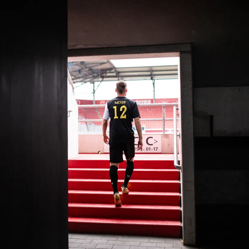 Een voetballer in een zwart shirt met nummer 12 en gele accenten, die de trappen naar een stadion afloopt.