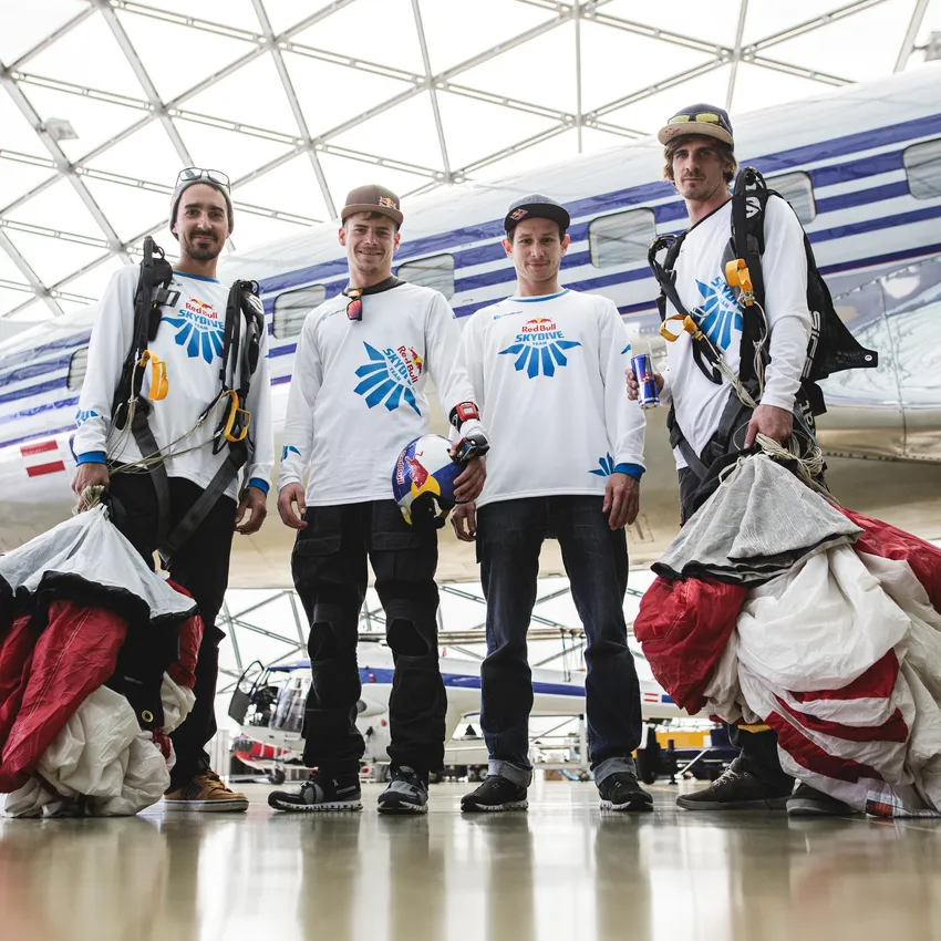 Vier mensen in sportkleding staan in een moderne hangar, klaar voor een parachutesprong.