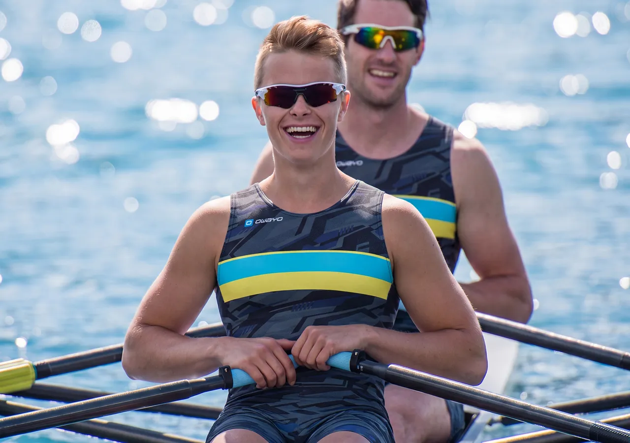 Twee atleten in stijlvolle sportkleding zitten in een roeiboot en glimlachen naar de camera terwijl ze peddelen op glinsterend water.