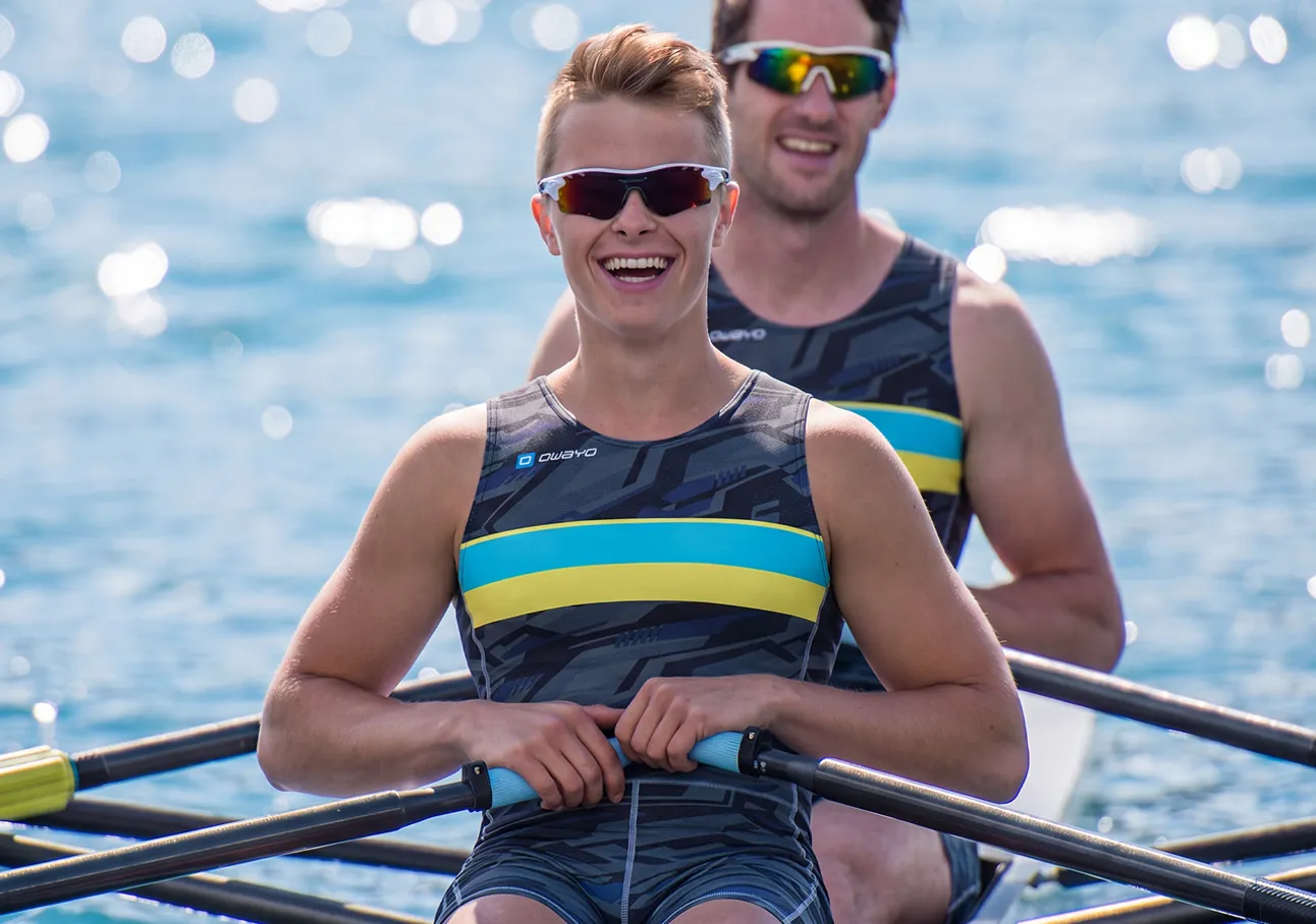 Twee blije roeiers in sportieve pakken met blauwe en gele strepen, zittend in een boot op sprankelend water.