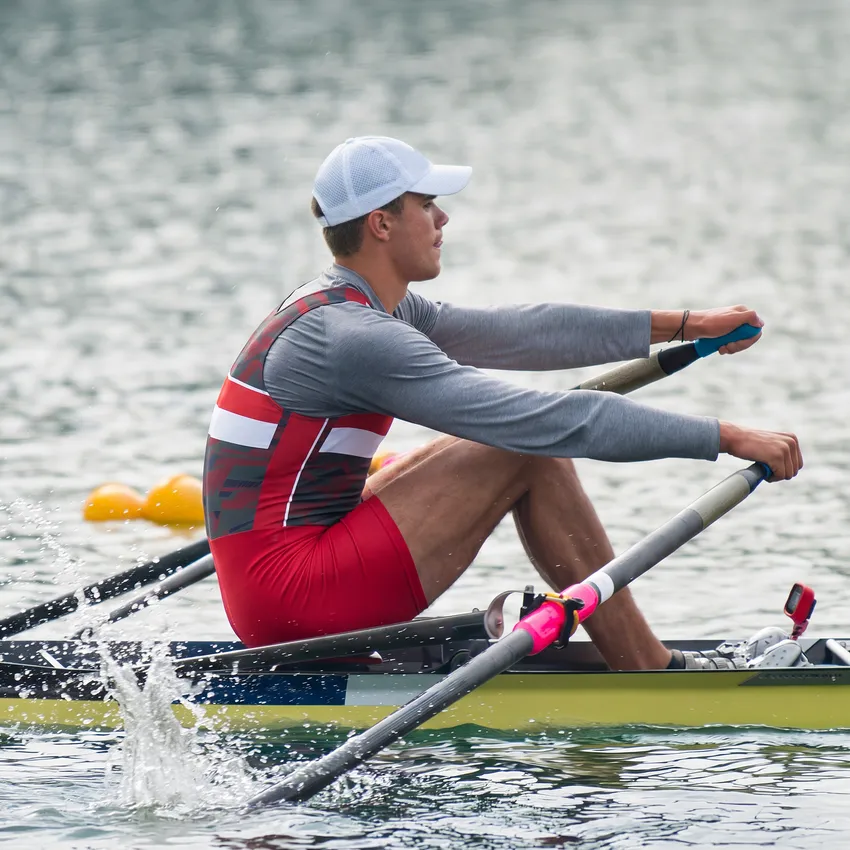 Een roeier in een rood en grijs sportoutfit roeit op een wateroppervlak.