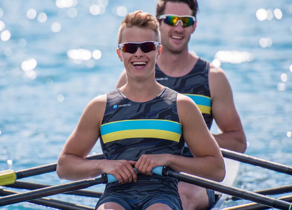 Twee roeiers in sportieve, gemusterde eenheden met blauwe en gele strepen, lachend op een wateroppervlak.