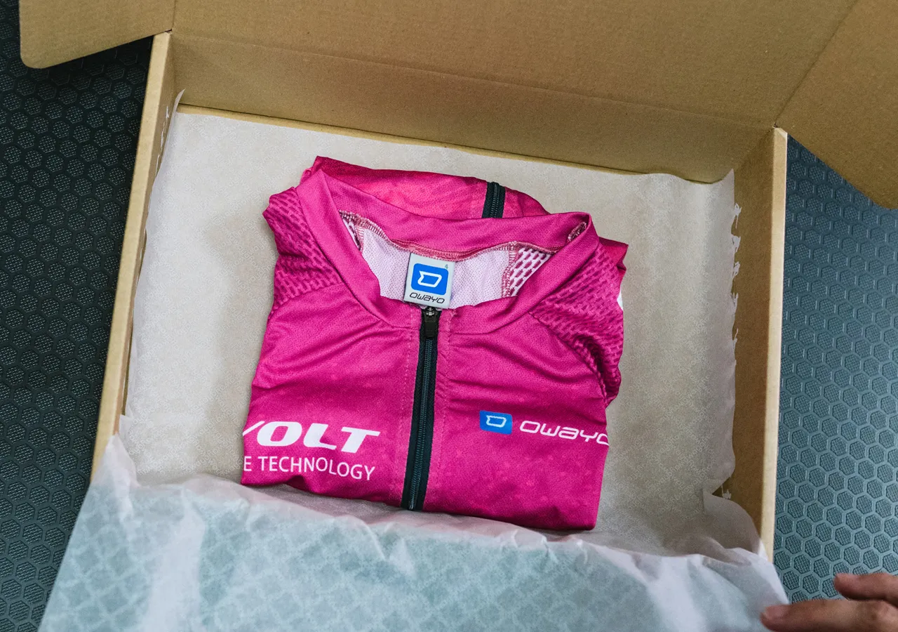 Een opgevouwen roze, mouwloos sportshirt met het owayo-logo ligt in een kartonnen doos op wit papier.
