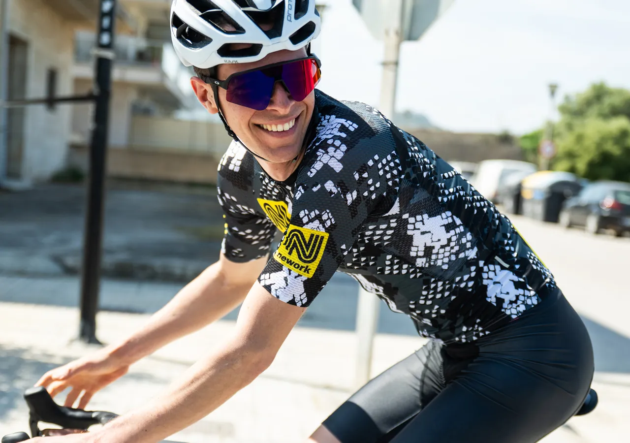 Een fietser in een zwart-wit gemusterd fietsshirt met een geel sponsorlogo, die op een straat fietst.