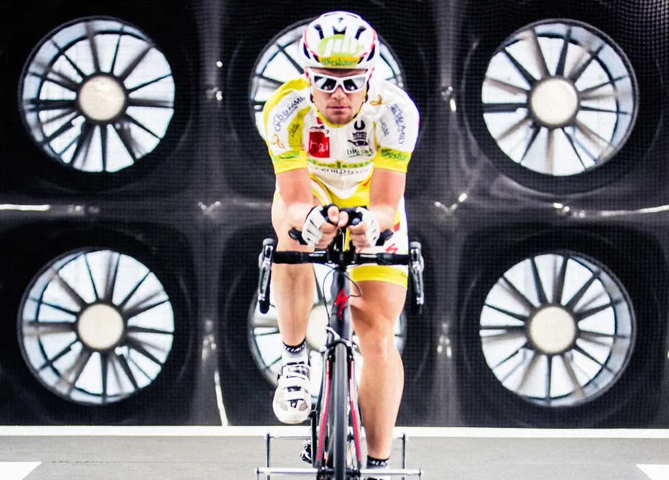 Een fietser in een geel-wit tricot en helm fietst op een fiets in een trainer voor een achtergrond met grote ventilatoren.