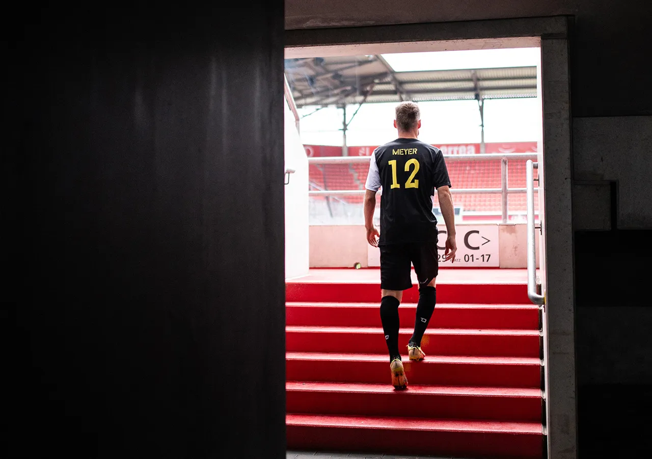 Een voetballer met de rugnummer 12 en de naam Meyer op het shirt loopt een rode trap op naar een stadion.