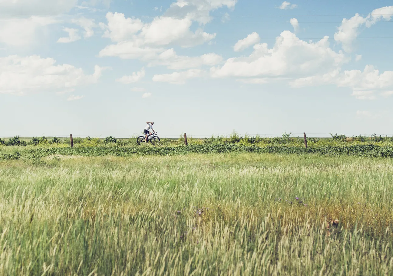 Een persoon fietst door een groen veld onder een blauwe lucht met witte wolken.