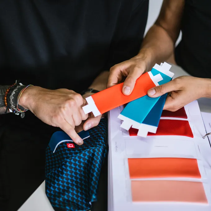 Twee personen kiezen kleurproeven voor aangepaste sportkleding, waarbij een blauwe stof met patroon en verschillende kleuropties zichtbaar zijn.